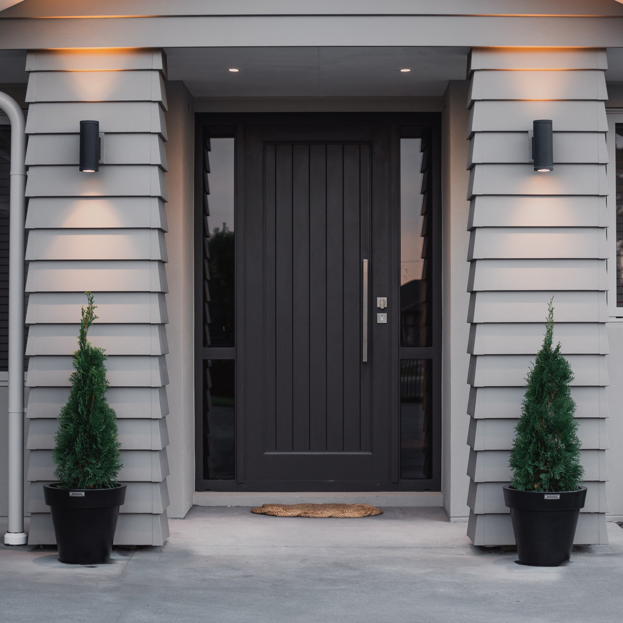 Two black planter pots either side of the front door of a house planted with Conifers.