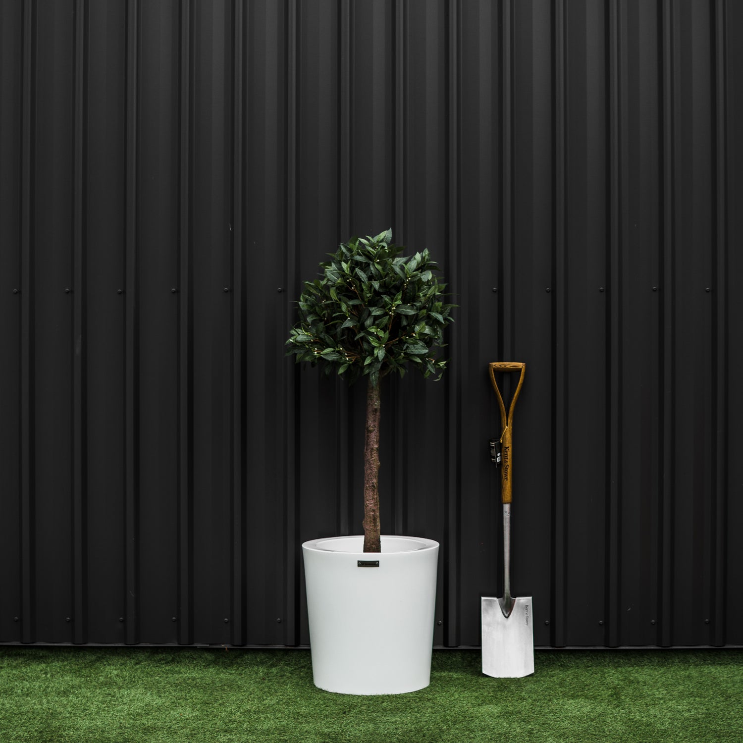 A white planter pot with a tree in it. The pot is beside a garden spade. Planter pots Australia.