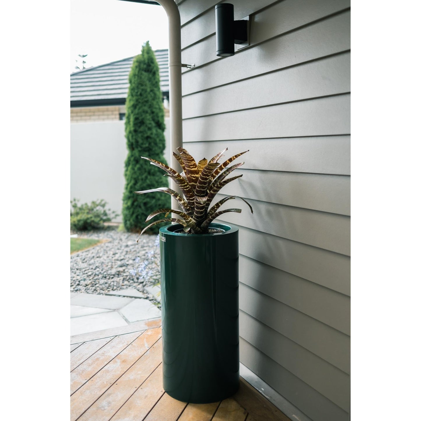 A cylinder planter pot sitting on a wooden deck beside a house.