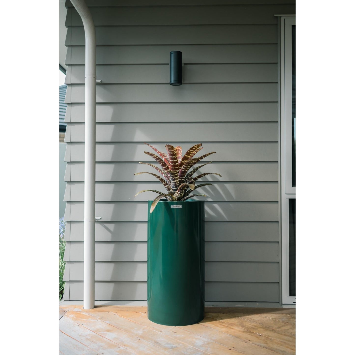 An emerald green cylinder planter pot sitting on a wooden deck in front of a house.
